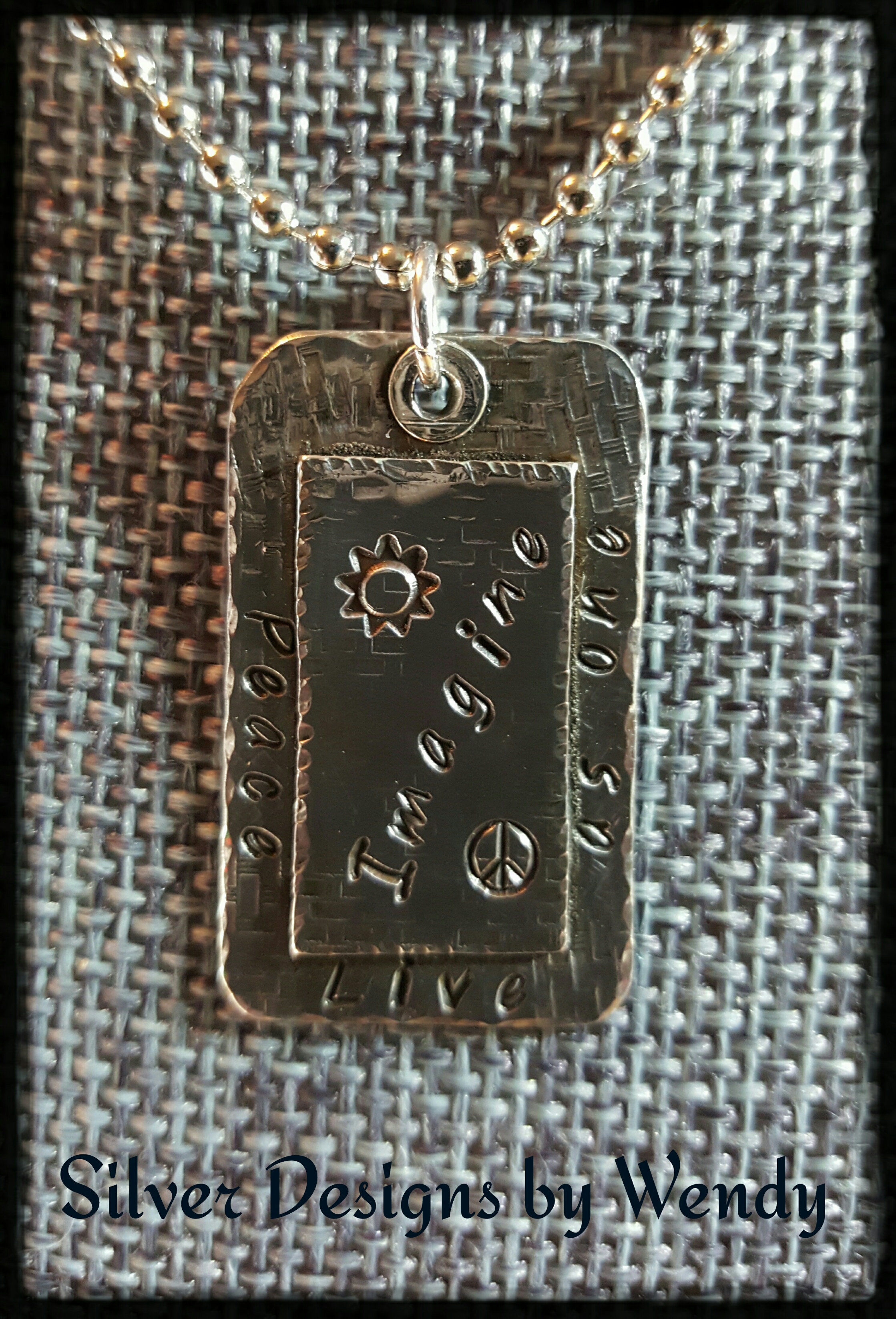 Silver necklace with the words peace, live as one, and imagine inscribed with a sun and peace sign