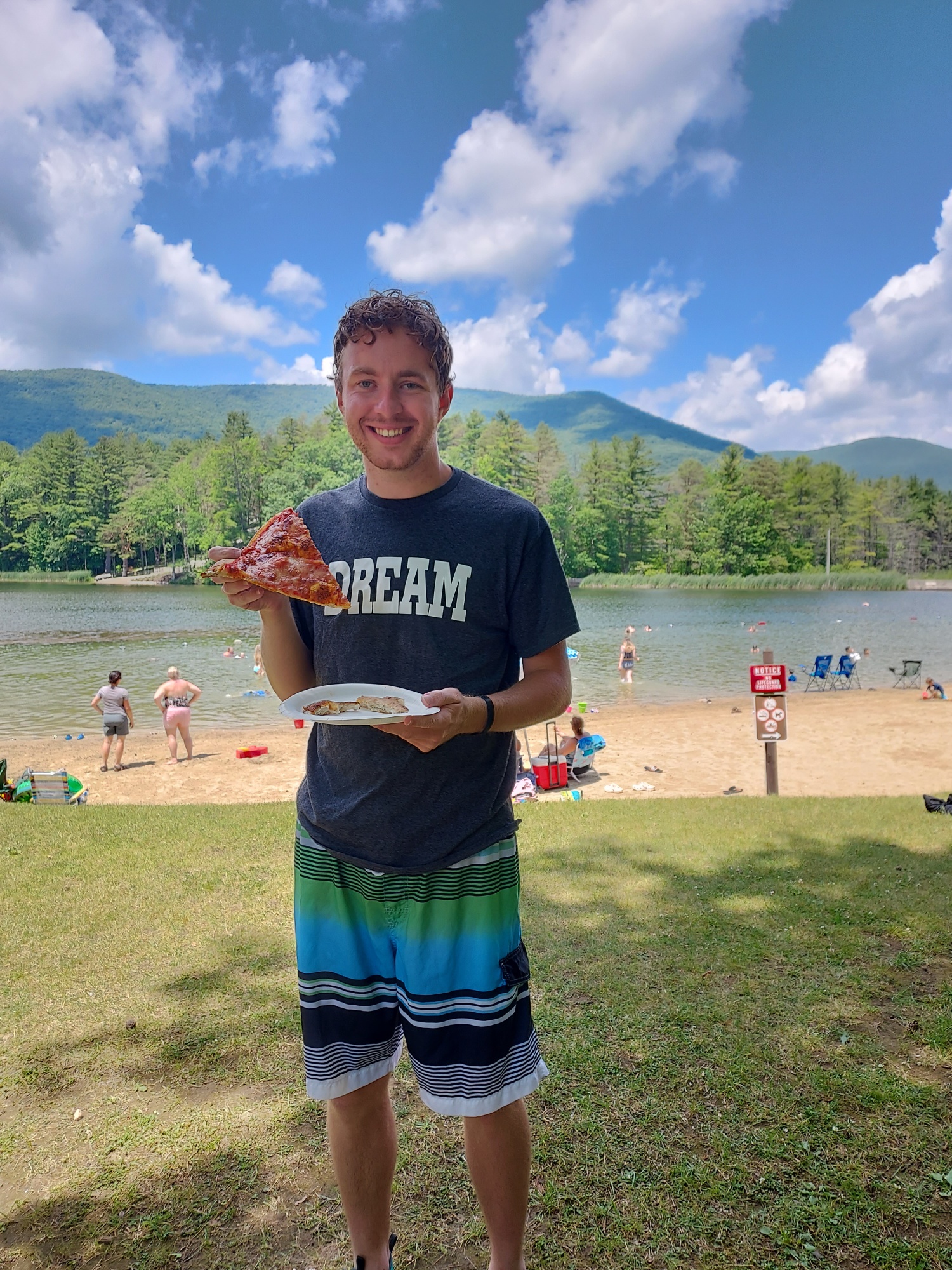 Clay holding a slice of pizza bigger than his head in front of a lake with a view of the forest and mountains.