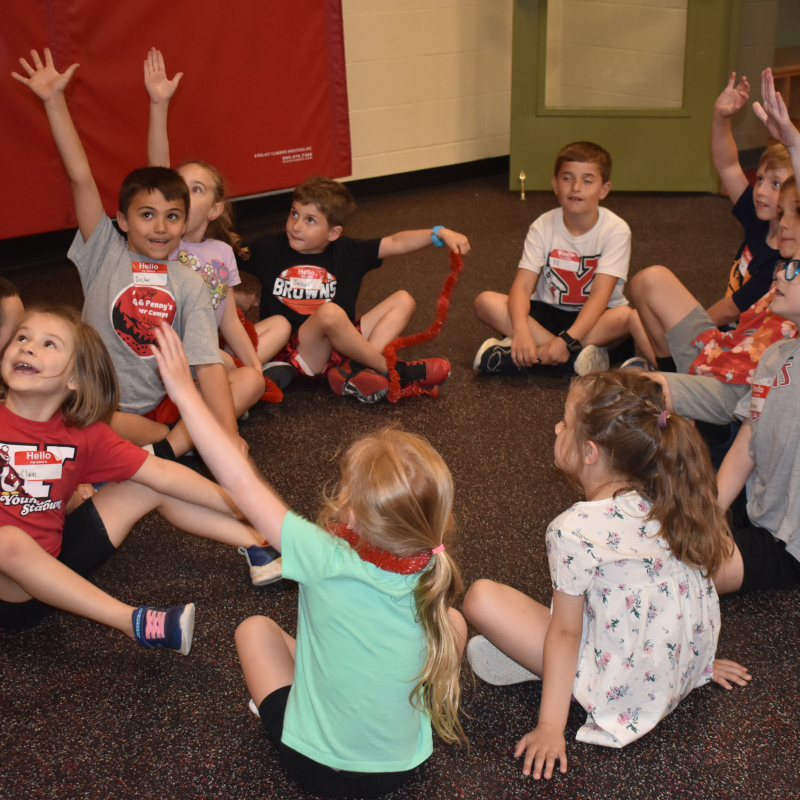 Group of children sitting on gym floor in a circle raising their hands.
