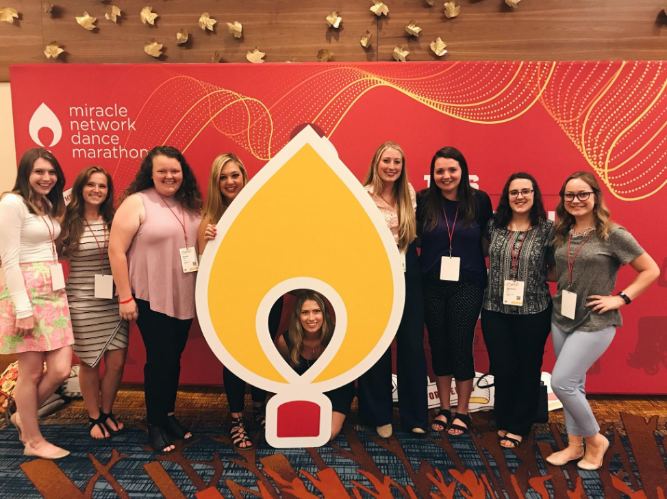 A group of students stand in front of the Dance Marathon logo and red and yellow background