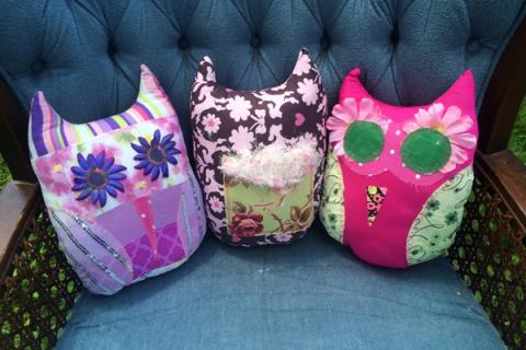 Three pillow owls propped up on a blue velvet chair. The owls range in a variety of colors between pink, brown, purple and green with flowers as eyes