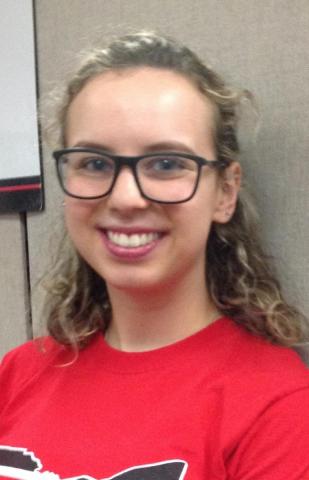 A smiling female student with glasses wearing a red shirt and posing against a wall