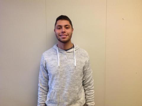 Smiling male student wearing a grey hooded sweatshirt posing against a wall