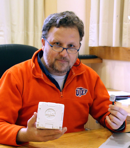 Male sitting at a desk looking directly at the camera, posing with equipment and wearing an orange pullover sweatshirt